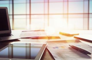 An office desk with financial reports, a touch pad, computer, pen and while the sun is shining in the office.