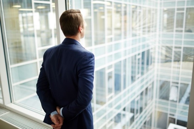 Rear view of elegant businessman standing by window of new office and looking through it