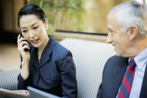 A businesswoman sitting on a couch in an office while talking on a phone and while a businessman looks at her and is smiling.