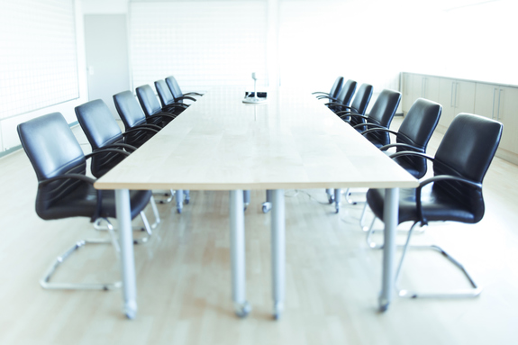 An image of a boardroom full of black chairs, a brown table and other items on the table.