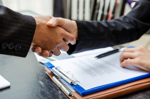 Two business people shaking hands in a office while one business person has their hand on a stack of documents on a table.