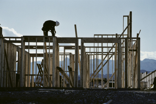 Construction worker with a white hard hat building the wooden frame of a house outside, with mountains, and the sky showing.