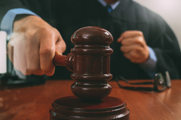 An image of a judge slamming a gavel on a sound block that is on a brown table with a pair of glasses on it.